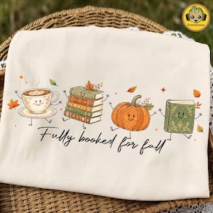 May include: A cream-colored t-shirt featuring a graphic print of a smiling teacup, a stack of books, an orange pumpkin, and a green book, all with arms and legs. The text "Fully booked for fall" is printed in black script below the illustrations.