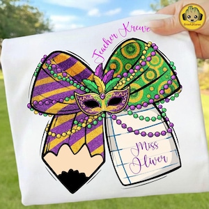 May include: White t-shirt featuring a colorful Mardi Gras design. The design includes a bow with purple and gold stripes, a mask, and a pencil. The text "Teacher Krewe" and "Miss Oliver" are also visible.
