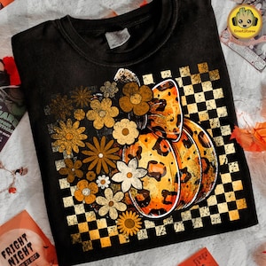May include: Black crew neck t-shirt featuring a pumpkin with a leopard print design and floral accents. The pumpkin is set against a checkered background with orange, white, and black squares. A fall-themed graphic tee.