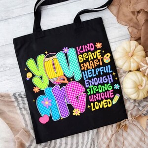 Op de afbeelding: Zwarte draagtas met de woorden "YOU ARE" in kleurrijke letters. De tas toont ook de woorden "KIND, BRAVE, SMART, HELPFUL, ENOUGH, STRONG, UNIQUE, LOVED". De tas heeft zwarte handvatten en is ontworpen voor het dragen van items.