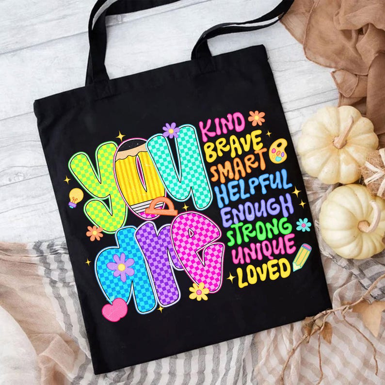 Op de afbeelding: Zwarte draagtas met de woorden "YOU ARE" in kleurrijke letters. De tas toont ook de woorden "KIND, BRAVE, SMART, HELPFUL, ENOUGH, STRONG, UNIQUE, LOVED". De tas heeft zwarte handvatten en is ontworpen voor het dragen van items.