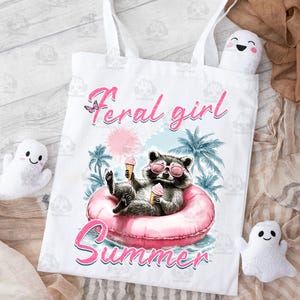 May include: White tote bag with a pink and blue illustration of a raccoon wearing sunglasses and holding an ice cream cone. The raccoon is sitting in a pink inflatable ring. The text "Feral girl summer" is printed in pink on the bag.
