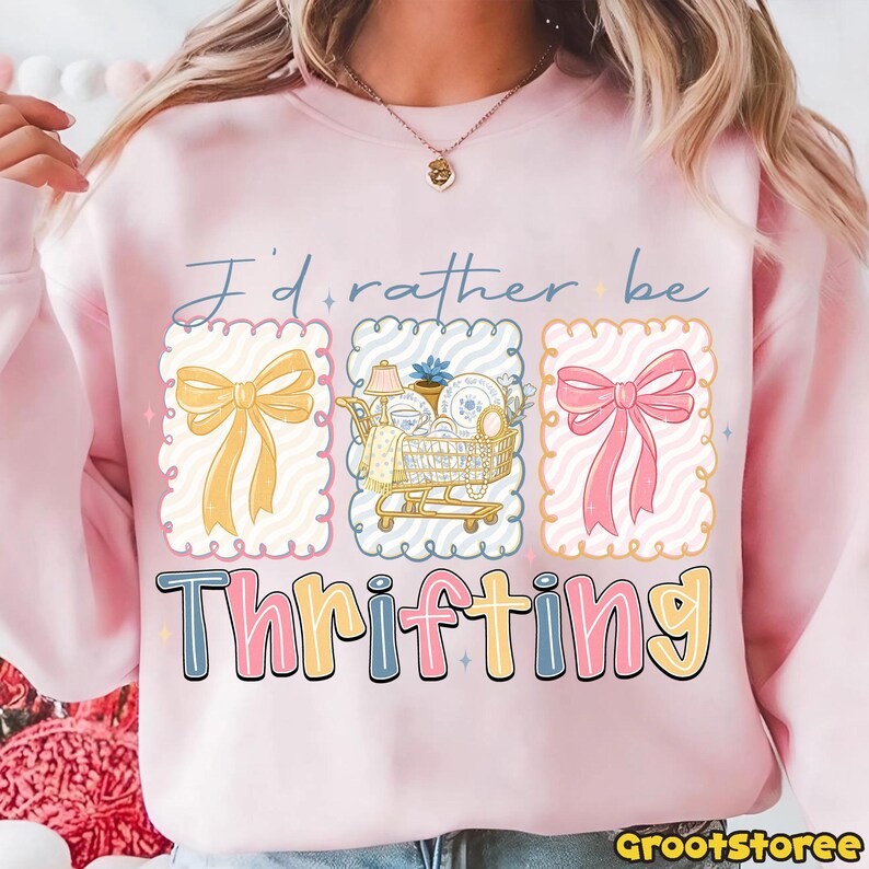 Może przedstawiać: R&oacute;żowa bluza z napisem "I'd rather be Thrifting". Projekt zawiera trzy oprawione ilustracje: ż&oacute;łtą kokardę, w&oacute;zek sklepowy wypełniony przedmiotami i r&oacute;żową kokardę. Słowo "Thrifting" jest w kolorowych literach.