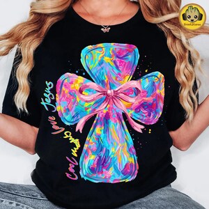 May include: Black t-shirt featuring a colorful cross design with a pink bow. The cross is painted with vibrant hues of blue, pink, yellow, and purple. The words "Love Jesus" and "Cool Vibes" are written in a cursive font on the shirt.
