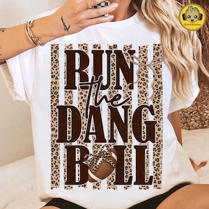 May include: A white t-shirt featuring a graphic design with vertical leopard print stripes, a brown bow, and a football. The text reads RUN The DANG BALL in large brown letters. The design is centered on the front of the shirt.