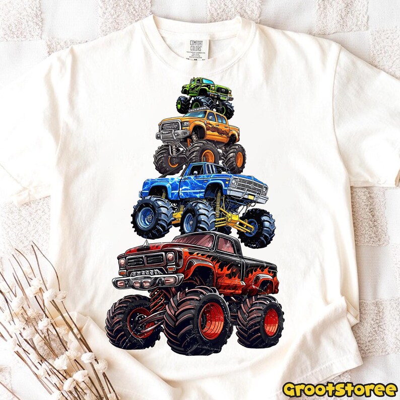 May include: White t-shirt featuring a graphic of five colorful monster trucks stacked on top of each other. The trucks are in shades of red, blue, orange, and green, with large tires. The text "GrootStoree" is at the bottom right.