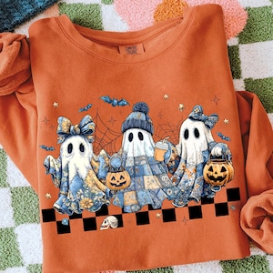 K&ouml;nnte beinhalten: Orangefarbenes Sweatshirt mit drei Cartoon-Geistern mit floralen und Patchwork-Designs, die K&uuml;rbislaternen und Getr&auml;nke halten. Die Geister sind mit Schleifen und H&uuml;ten verziert, mit Flederm&auml;usen und Sternen im Hintergrund. Das Shirt hat ein Comfort Colors-Etikett.