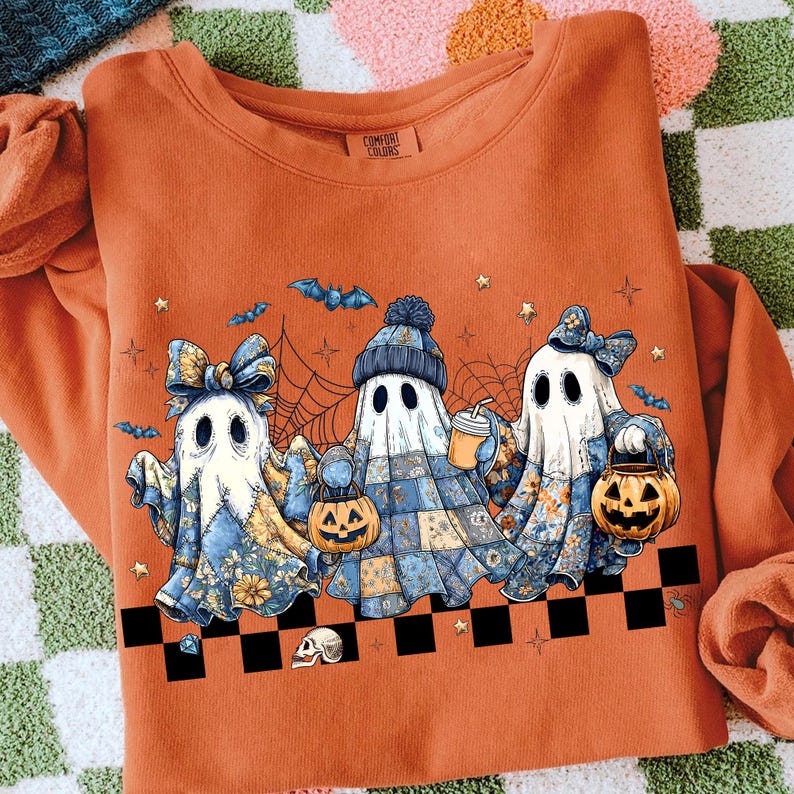 K&ouml;nnte beinhalten: Orangefarbenes Sweatshirt mit drei Cartoon-Geistern mit floralen und Patchwork-Designs, die K&uuml;rbislaternen und Getr&auml;nke halten. Die Geister sind mit Schleifen und H&uuml;ten verziert, mit Flederm&auml;usen und Sternen im Hintergrund. Das Shirt hat ein Comfort Colors-Etikett.