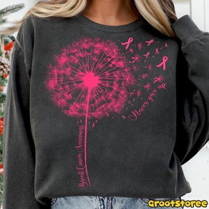 May include: Dark gray sweatshirt featuring a pink dandelion design with the words "Breast Cancer Awareness" and "Never give up." Pink ribbons are scattered around the dandelion. The sweatshirt has a crew neck and long sleeves.