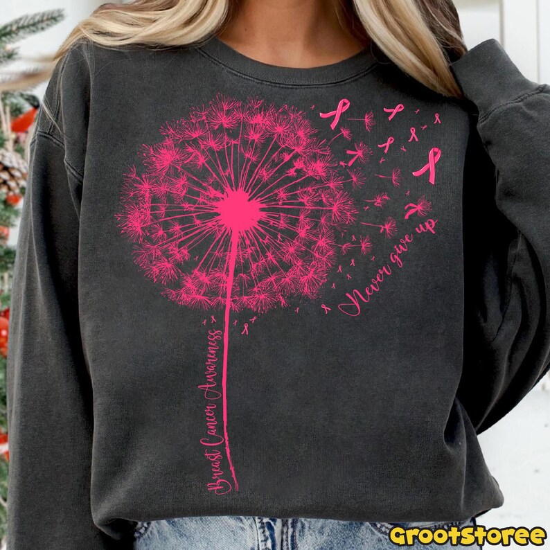 May include: Dark gray sweatshirt featuring a pink dandelion design with the words "Breast Cancer Awareness" and "Never give up." Pink ribbons are scattered around the dandelion. The sweatshirt has a crew neck and long sleeves.