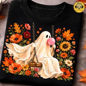 May include: A black t-shirt featuring a graphic of a white ghost sitting on a wooden swing, blowing a pink bubble with bubblegum. The ghost is surrounded by vibrant autumn-colored flowers and leaves, including orange poppies and sunflowers.