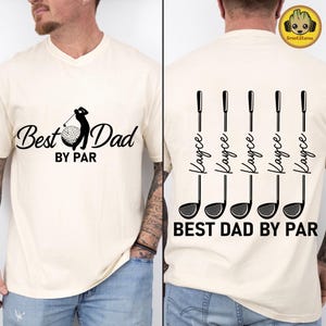 May include: A cream-colored t-shirt featuring a black graphic on the front with a golfer silhouette, a golf ball, and the text "Best Dad BY PAR". The back displays five vertical golf club illustrations with the name "Kayce" and "BEST DAD BY PAR".