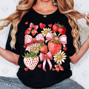 May include: A black t-shirt with a pink and red strawberry and bow design. The design features strawberries, bows, hearts, and daisies.