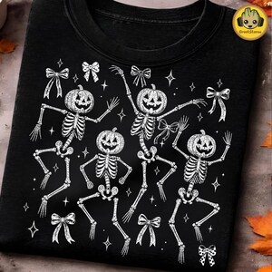 May include: A black t-shirt featuring a white graphic print of four skeletons with jack-o'-lantern heads in various dancing poses. The design is accented with multiple small bows and four-pointed stars scattered across the fabric surface.