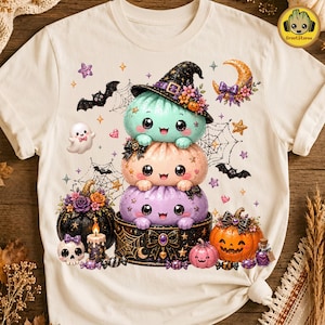 May include: A cream-colored t-shirt featuring a graphic of three stacked, smiling, round creatures in mint, peach, and purple. The top creature wears a black witch hat. Surrounding them are pumpkins, a ghost, bats, spiderwebs, and a candle.