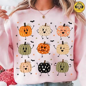 May include: A light pink crewneck sweatshirt featuring a graphic print of nine cartoon pumpkins with various facial expressions, patterns, and colors, interspersed with small orange stars and black bat silhouettes across the front.