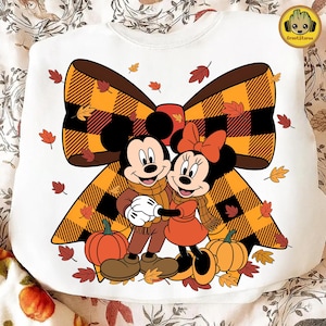 May include: White sweatshirt featuring Mickey and Minnie Mouse embracing, with a large plaid bow and autumn leaves. Mickey wears a brown outfit and scarf, Minnie a dress and bow in orange. Pumpkins are at the bottom. The GrootStore logo is in the top right.