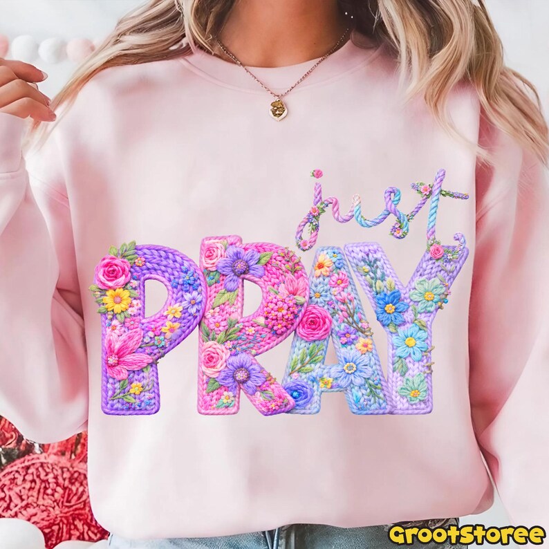 May include: A light pink sweatshirt with the words "just PRAY" in a floral design. The letters are in shades of purple, pink, and blue, with flowers and leaves. The word "just" is in a script font.