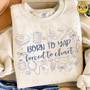 May include: Cream-colored sweatshirt with blue medical-themed illustrations and the text "BORN TO YAP forced to chart." The design includes a stethoscope, syringe, and other medical symbols. A comfortable, casual top.
