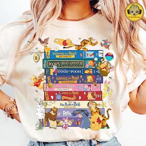 May include: Cream-colored t-shirt featuring a stack of Winnie the Pooh VHS tapes with various characters. The design includes bees, flowers, and the text "Winnie the Pooh" on several tapes. The Groot Storee logo is in the upper right corner.