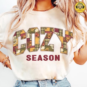 Op de afbeelding: Crèmekleurig T-shirt met de tekst "COZY SEASON" in een patchwork-ontwerp. De letters zijn gevuld met bloemen- en ruitpatronen in tinten oranje, groen en bordeaux. De tekst is in een schreefletter.