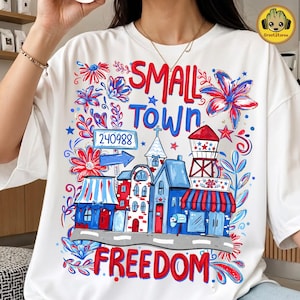 Puede incluir: Camiseta blanca con un diseño gráfico colorido. El diseño presenta las palabras "SMALL TOWN FREEDOM" en rojo y azul, junto con ilustraciones de edificios, flores y estrellas. Una flecha azul apunta al número 240988.