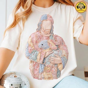Könnte beinhalten: Cremefarbenes T-Shirt mit einem Patchwork-Design, das eine Figur zeigt, die einen blau-weiß karierten Hasen hält. Der Patchwork-Stoff enthält Blumen- und Tupfenmuster in Pastellfarben. Das Design ist mittig auf dem Shirt platziert.