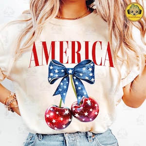 May include: White t-shirt with the word "AMERICA" printed in red letters. A blue and white bow with stars is tied around two red cherries with white stars.