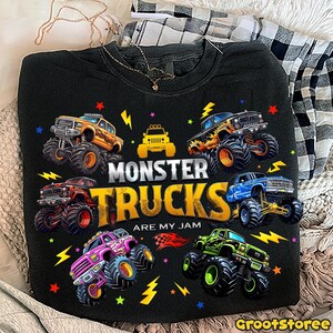 May include: Black sweatshirt with a colorful graphic of monster trucks. The design includes the text "MONSTER TRUCKS ARE MY JAM" with various monster truck illustrations in orange, red, blue, green, and pink. The design also includes lightning bolts and stars.