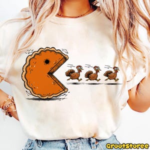 May include: A cream-colored t-shirt featuring a humorous Thanksgiving design. The graphic depicts a pie slice in the style of Pac-Man chasing three cartoon turkeys wearing pilgrim hats. The text "GrootStoree" is printed in the bottom right corner.