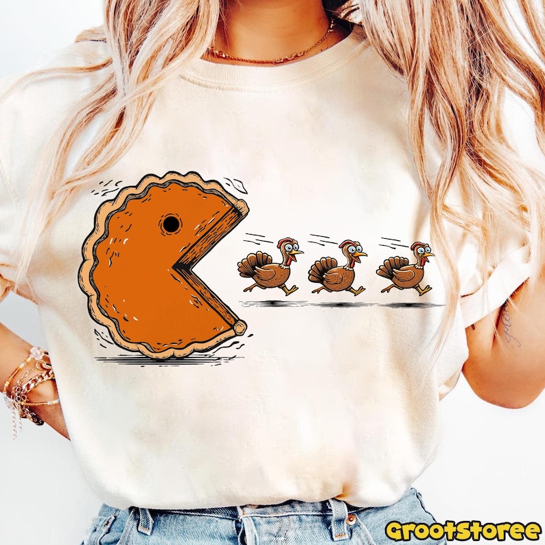 May include: A cream-colored t-shirt featuring a humorous Thanksgiving design. The graphic depicts a pie slice in the style of Pac-Man chasing three cartoon turkeys wearing pilgrim hats. The text "GrootStoree" is printed in the bottom right corner.