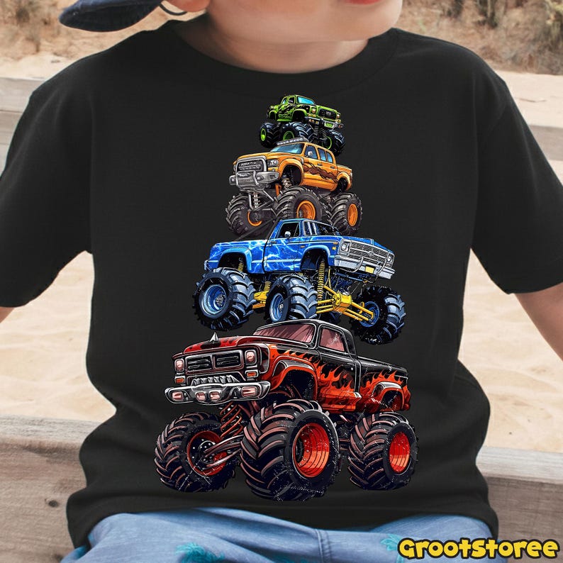 May include: Black t-shirt featuring a stack of colorful monster trucks. The trucks are red, orange, blue, and green, with large tires and detailed graphics. The text "GrootStoree" is at the bottom right.