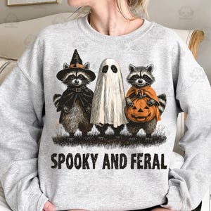 May include: A light gray sweatshirt featuring a Halloween design. The graphic includes three raccoons dressed as a witch, a ghost, and a pumpkin. The text "SPOOKY AND FERAL" is printed in black below the characters. The design is centered on the front of the sweatshirt.