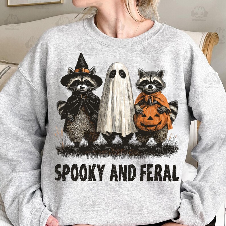May include: A light gray sweatshirt featuring a Halloween design. The graphic includes three raccoons dressed as a witch, a ghost, and a pumpkin. The text "SPOOKY AND FERAL" is printed in black below the characters. The design is centered on the front of the sweatshirt.