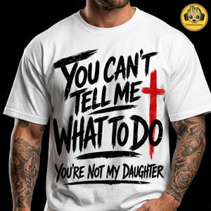 Puede incluir: Una camiseta blanca de manga corta con letras negras desgastadas que dicen "YOU CAN'T TELL ME WHAT TO DO" y "YOU'RE NOT MY DAUGHTER", junto a un símbolo de cruz roja prominente integrado en el lado derecho del diseño.