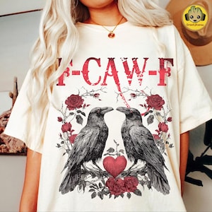 Puede incluir: Camiseta color crema con un diseño gráfico. El diseño presenta el texto "F-CAW-F" en rojo, dos cuervos negros, rosas rojas y un corazón. Una prenda casual para el uso diario.