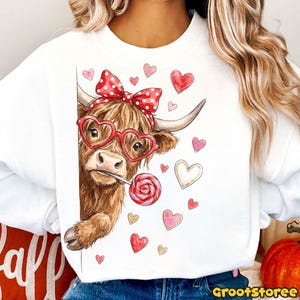 May include: White sweatshirt featuring a brown cow with heart-shaped glasses and a red polka dot bow. The cow is holding a lollipop and surrounded by pink and gold hearts. The sweatshirt has a crew neck.