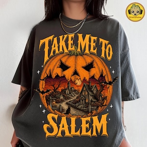 Pode incluir: Uma t-shirt cinzento-carvão com um gráfico de uma grande abóbora laranja contendo uma cena de uma aldeia assustadora, um gato preto e morcegos. O texto "TAKE ME TO SALEM" está impresso em letras laranja acima e abaixo da abóbora.