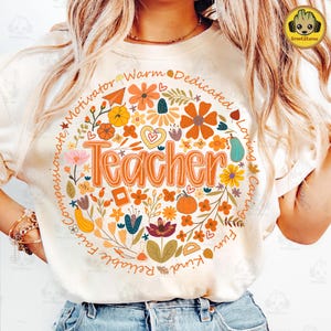 May include: Cream-colored t-shirt with a floral design surrounding the word "Teacher" in orange. The design includes flowers, leaves, and the words "Motivator", "Warm", "Dedicated", "Loving", "Caring", "Fun", "Kind", "Reliable", "Fair", and "Compassionate".