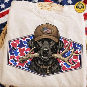 Può includere: Una felpa beige con un Labrador Retriever nero che indossa un berretto marrone con una bandiera americana. Il cane tiene le corna in bocca, su un design mimetico e geometrico. Un'etichetta sul collare recita "1776".