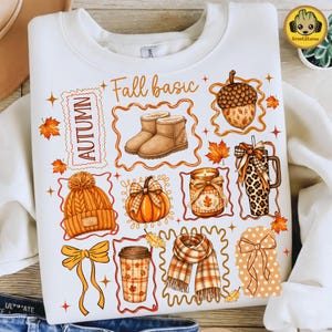 May include: A white crewneck sweatshirt featuring a colourful graphic print of autumn-themed items including tan boots, an acorn, a pumpkin, a knit hat, a plaid scarf, a coffee cup, and a tumbler, with the text "AUTUMN" and "Fall basic" in orange.