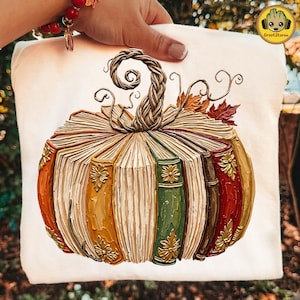 Peut inclure: Un t-shirt blanc avec une impression graphique repr&eacute;sentant une citrouille form&eacute;e par des livres anciens empil&eacute;s. La citrouille poss&egrave;de une tige tress&eacute;e, des vrilles et des feuilles d'automne, avec des dos de livres orange, verts, marron et rouges.