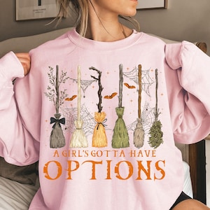 May include: Pink sweatshirt with a graphic of several witch brooms in various styles, with spiderwebs and bats. The text "A GIRL'S GOTTA HAVE OPTIONS" is printed in orange below the brooms. A casual, Halloween-themed design.