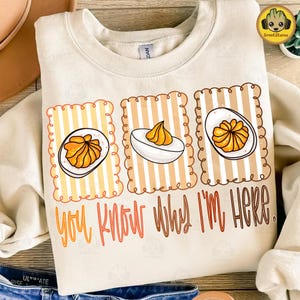 Può includere: Felpa color crema con una grafica di uova ripiene in una cornice a righe. Il testo "You Know Why I'm Here" è stampato in arancione e marrone. Il design include tre illustrazioni di uova ripiene.