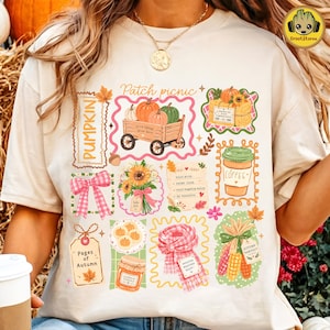May include: A cream-colored t-shirt featuring a graphic print with the text Patch picnic, Pumpkin, Pages of Autumn, and Sweater Weather. The design includes illustrations of a pumpkin wagon, sunflowers, a coffee cup, a scarf, and autumn treats.