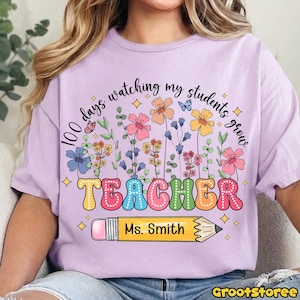 May include: A light purple t-shirt with the text "100 days watching my students grow" above a colorful floral design and the word "TEACHER" in large, bubble letters. A yellow pencil with the name "Ms. Smith" is below the word "TEACHER".