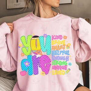 Op de afbeelding: Een lichtroze sweatshirt met de woorden "YOU ARE" in grote, kleurrijke letters. De woorden "KIND, BRAVE, SMART, HELPFUL, ENOUGH, STRONG, UNIQUE, LOVED" zijn kleiner geschreven. Het ontwerp bevat bloemen, harten en een potlood.