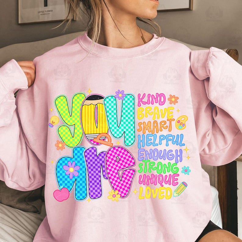 Op de afbeelding: Een lichtroze sweatshirt met de woorden "YOU ARE" in grote, kleurrijke letters. De woorden "KIND, BRAVE, SMART, HELPFUL, ENOUGH, STRONG, UNIQUE, LOVED" zijn kleiner geschreven. Het ontwerp bevat bloemen, harten en een potlood.