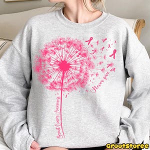 May include: A light gray sweatshirt featuring a pink dandelion design with the words "Breast Cancer Awareness" and "Never give up". The dandelion's seeds are replaced with pink ribbons, symbolizing breast cancer awareness.