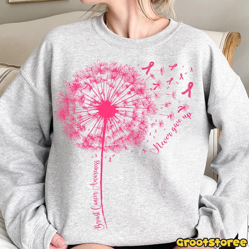 May include: A light gray sweatshirt featuring a pink dandelion design with the words "Breast Cancer Awareness" and "Never give up". The dandelion's seeds are replaced with pink ribbons, symbolizing breast cancer awareness.